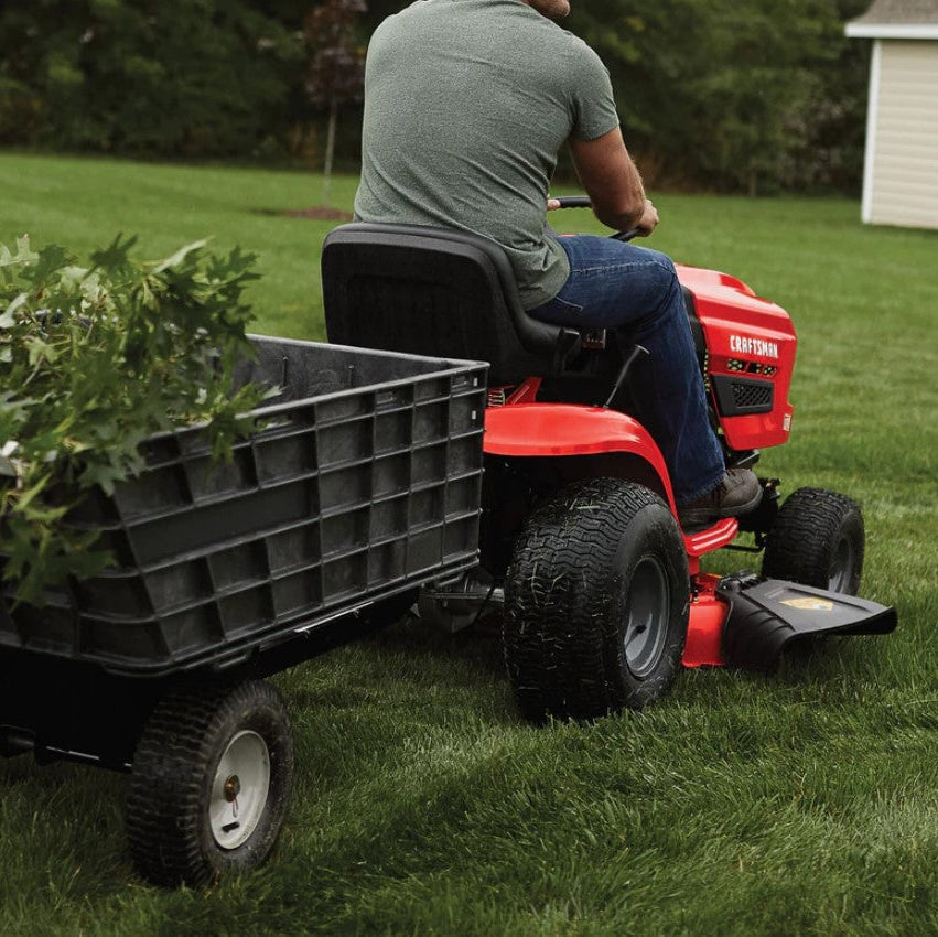 Person using a red riding lawn mower with a black trailer carrying plants on a grassy area.